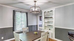 Un coin repas comprenant une table en bois, des chaises blanches, un lustre et une étagère intégrée où sont rangés des verres.