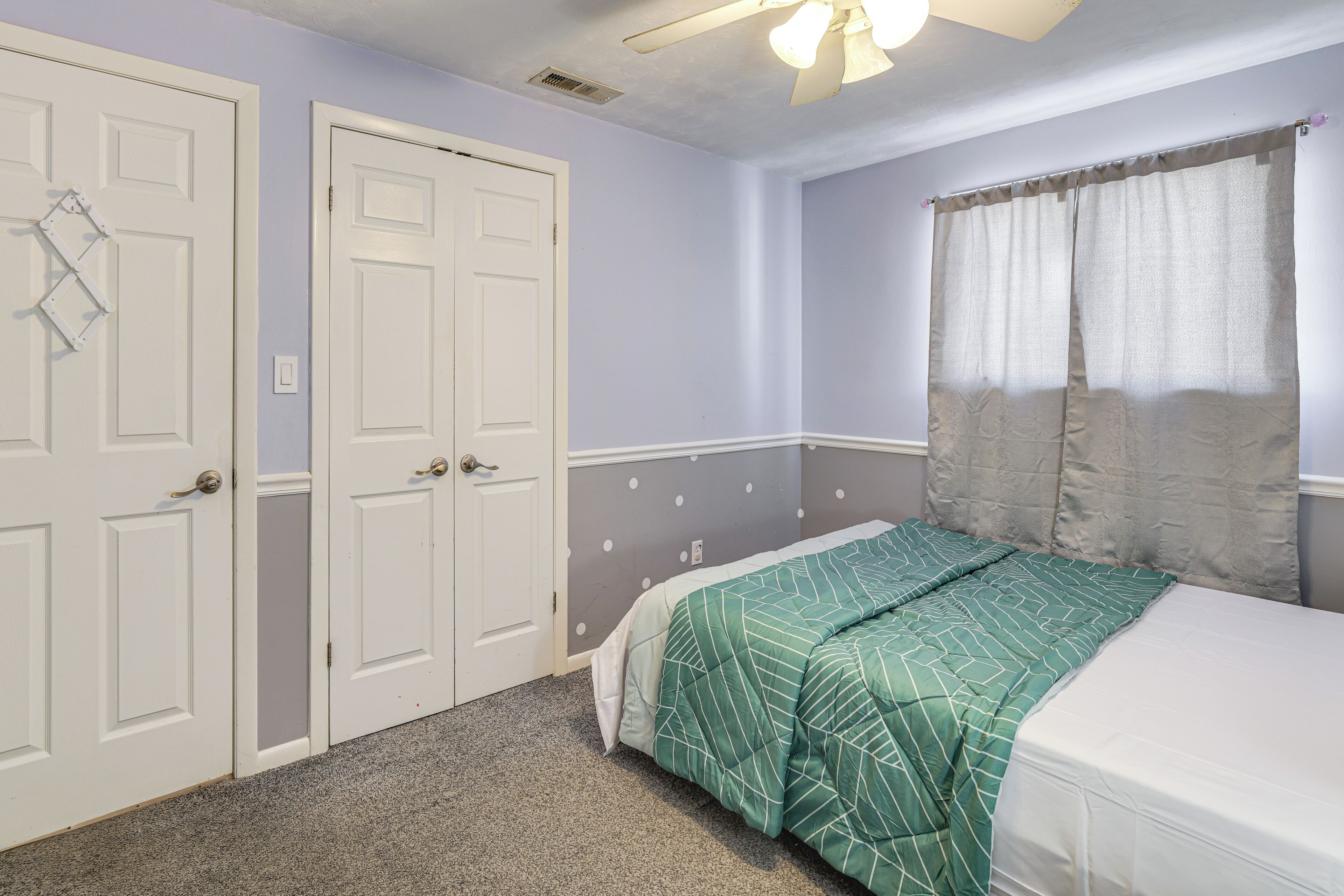 Un dormitorio con cama, dos puertas blancas, ventilador de techo y una ventana con cortinas.