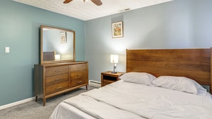 Een slaapkamer met een houten hoofdbord, een groot bed, een dressoir met lades, een nachtkastje met een lamp en een spiegel aan de muur.
