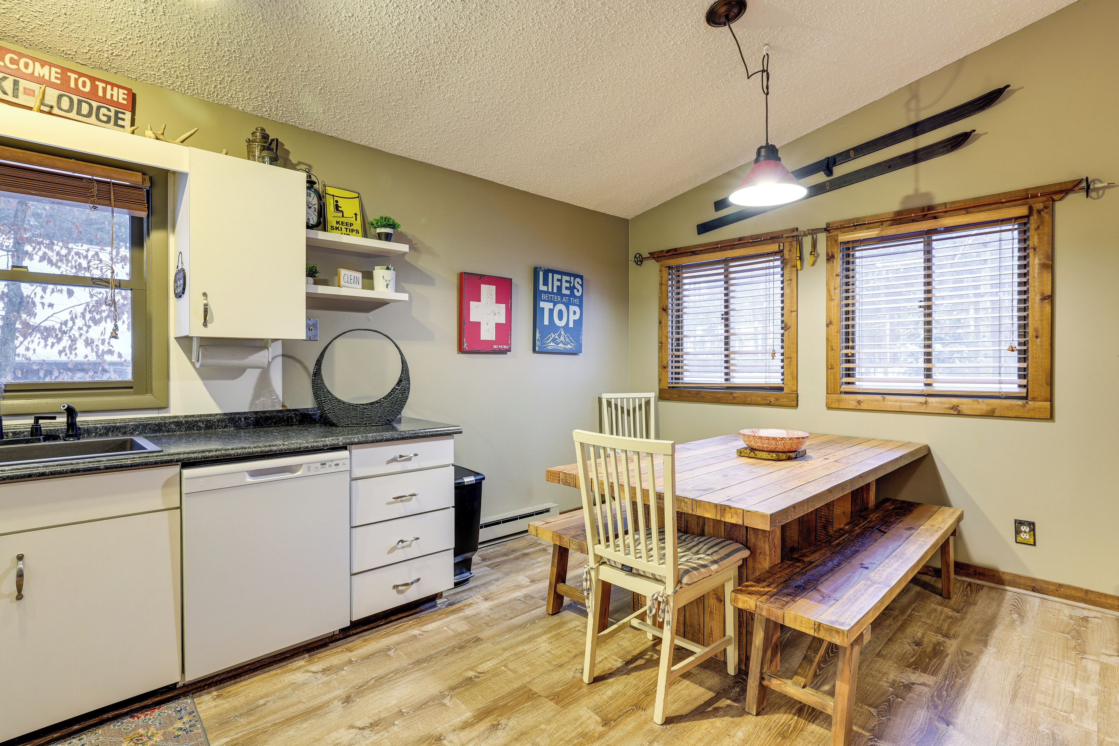 Una cocina con armarios blancos, una mesa de comedor de madera y un banco. Hay dos ventanas con persianas, un ventilador de techo y un cartel que dice "Welcome to the Lodge".
