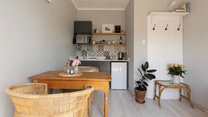 A compact kitchen with a dining area, featuring a wooden table, wicker chairs, and a small side table with a potted plant. - SurfWay beach cottage - Your Cape beach getaway (Cape Town)