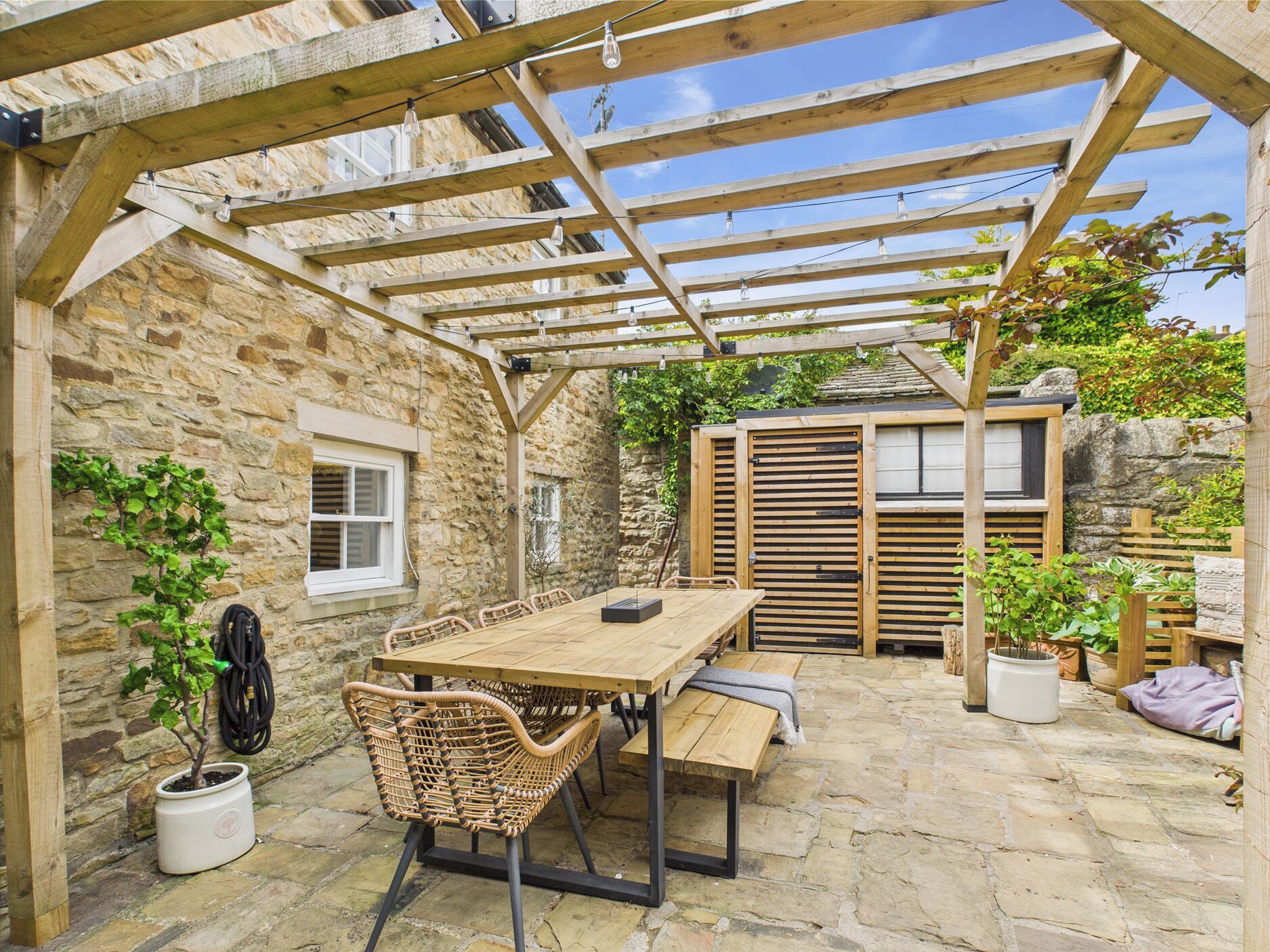 Un patio de paredes de piedra con una pérgola de madera, una mesa con sillas y plantas en macetas.