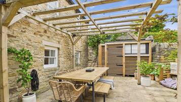 Un patio de paredes de piedra con una pérgola de madera, una mesa con sillas y plantas en macetas.