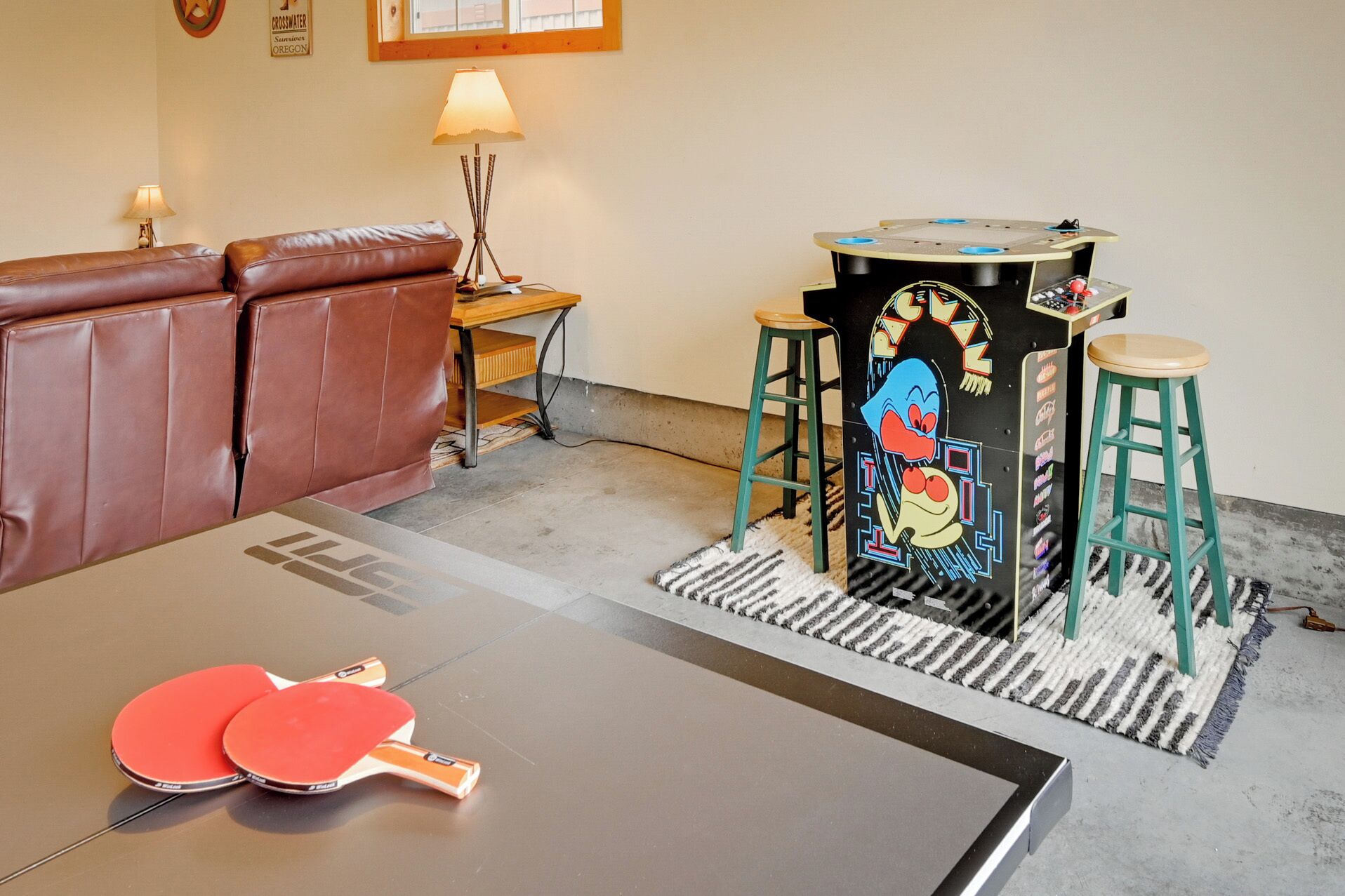 Et ping pong-bord med to ketchere, en lædersofa i brunt, en lampe og en farverig arkademaskine.