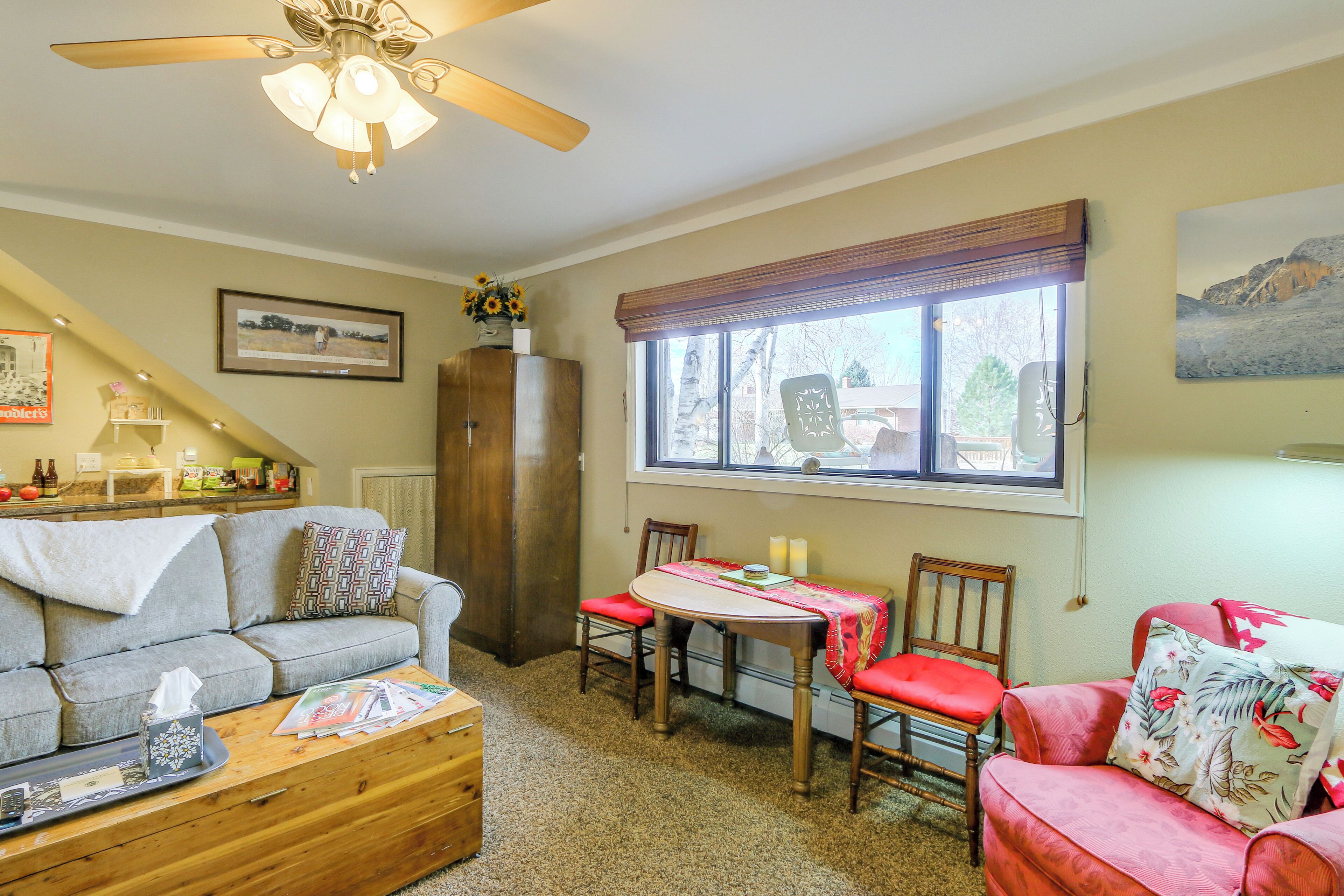 A cozy living room with a sofa, a dining table, and chairs. There is a ceiling fan, a window with blinds, and a painting on the wall.