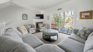 Ein Wohnzimmer mit grauem Ecksofa, einem runden Hocker, einem Kamin, einem Fernseher und einer großen Glasschiebetür, die zu einem Außenbereich führt.
