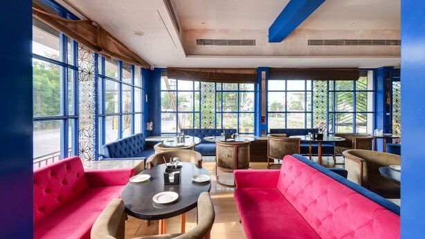 A restaurant with large windows, red and blue upholstered seating, and multiple tables. - Hotel Maanvad Garh by Keshav Residency (Udaipur)