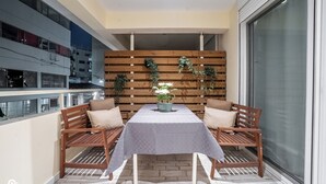 Ein Balkon mit Tisch und zwei Holzsesseln, eine Holzverkleidung mit hängenden Pflanzen und ein Blick auf ein Gebäude im Hintergrund.