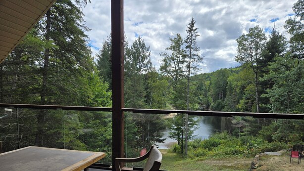 Una mesa de madera y dos sillas en un balcón con vista a un lago rodeado de árboles.