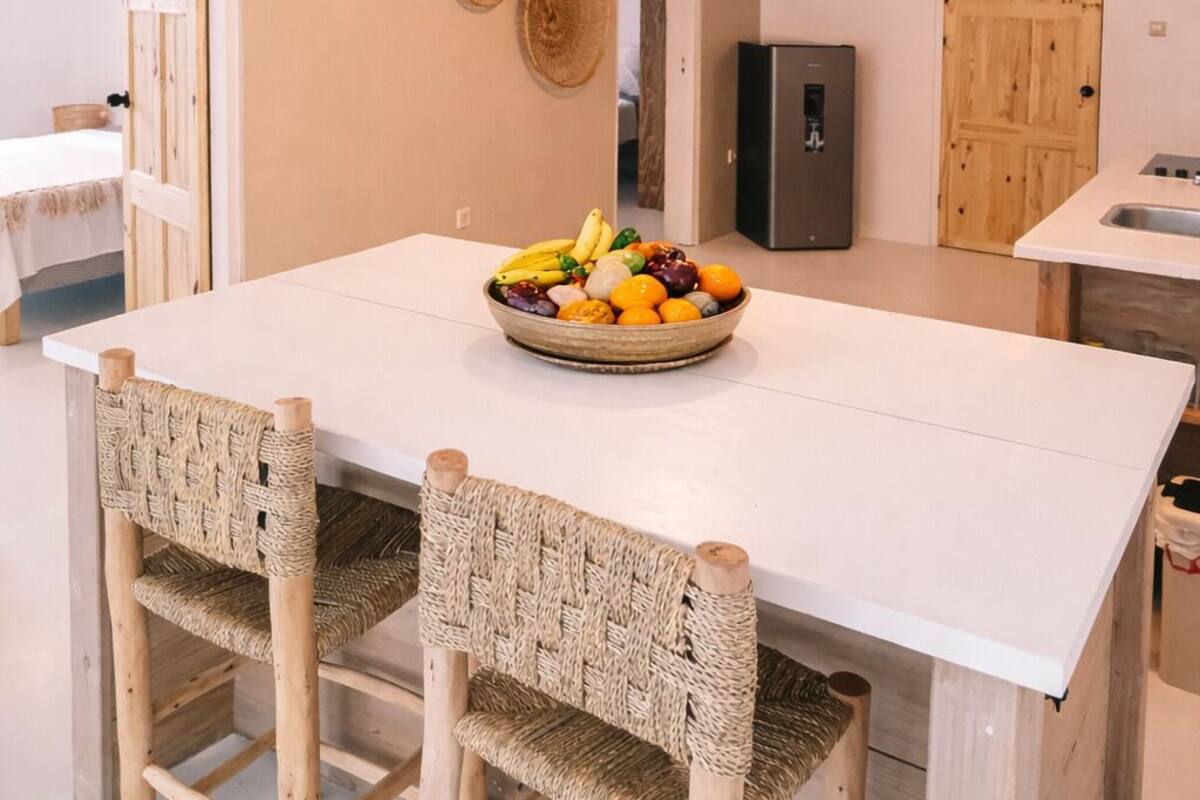 Une cuisine moderne dotée d’un grand îlot central, de chaises en osier, d’un panier de fruits et d’une porte en bois.