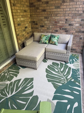 A wicker sofa with cushions against a brick wall, a green and white patterned rug, and a green door. - Beautiful 1st Floor, Pet Friendly Condo in Barefoot Landing (North Myrtle Beach)