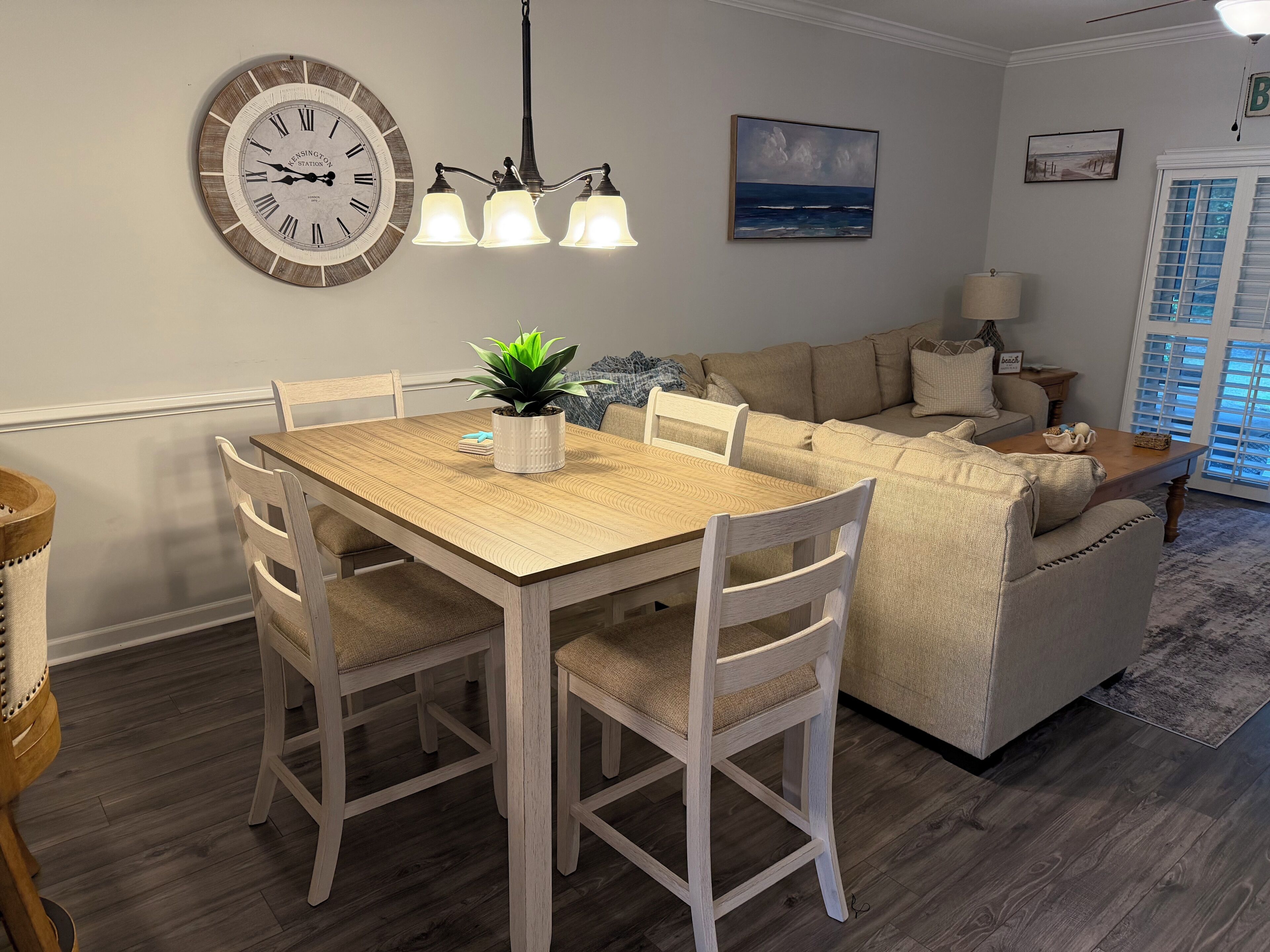 Un coin repas avec une table et des chaises en bois, une horloge murale, un lustre, et un salon avec un canapé et un tableau.