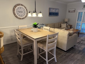 Un coin repas avec une table et des chaises en bois, une horloge murale, un lustre, et un salon avec un canapé et un tableau.