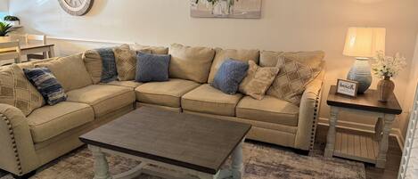 A living room with a beige sectional sofa, a wooden coffee table, a rug, a wall clock, a floral painting, a lamp, and a small table with a sign.