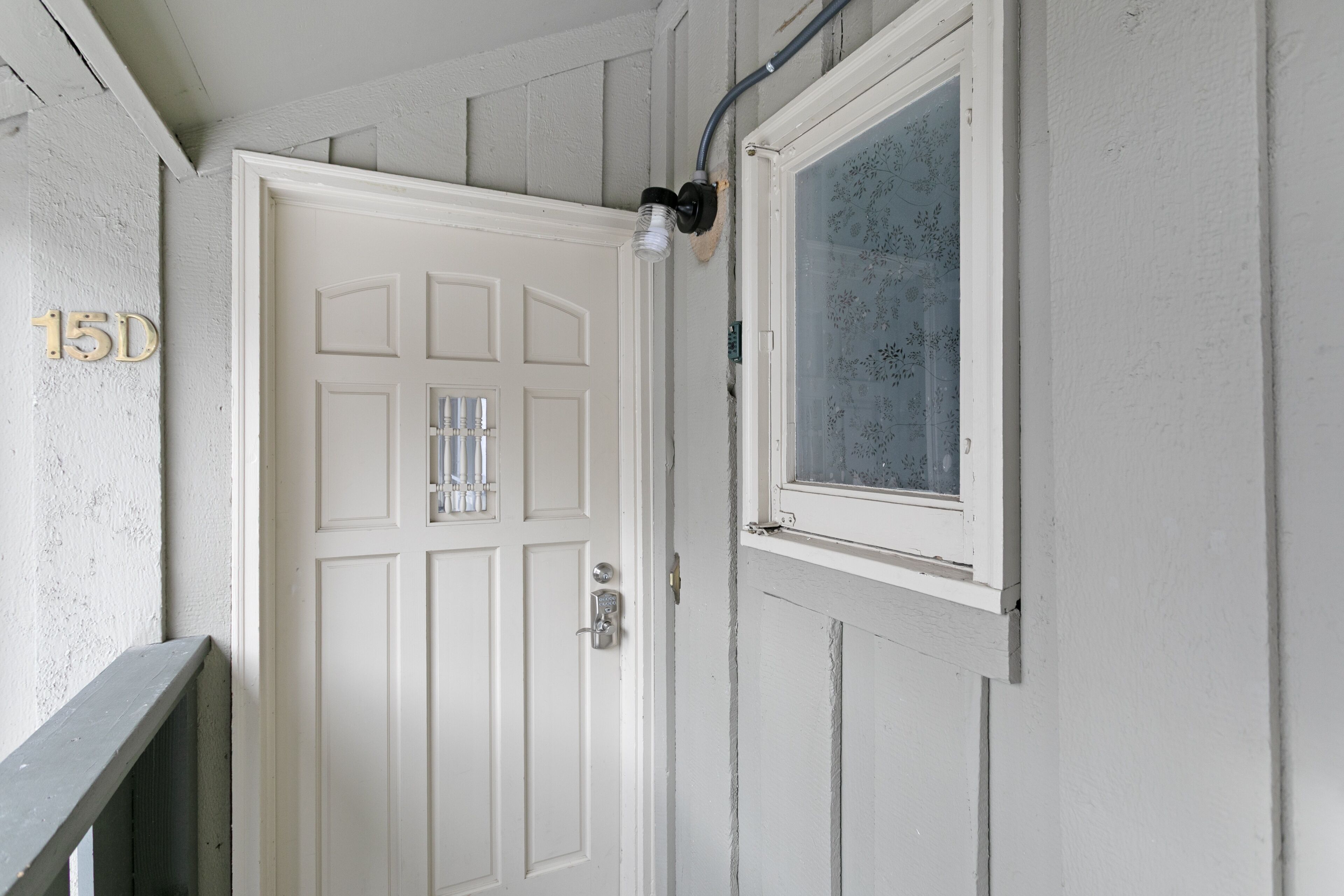 Une porte blanche avec une fenêtre, une petite fenêtre ornée d’une grille décorative, et le numéro « 15D » peint sur le mur.