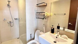 A modern bathroom with a glass shower enclosure, a toilet, a sink with a mirror, and a small shelf with toiletries. - Room-stylish Studio Stay at Saint Domini (Iloilo)