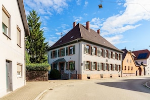 Un bâtiment à deux étages de style architectural européen traditionnel, caractérisé par une façade symétrique, de nombreuses fenêtres avec volets et une entrée située à l’angle.