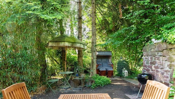 Een houten tafel met stoelen in een tuin, met een kiosk, een stenen muur en een barbecueplaats.