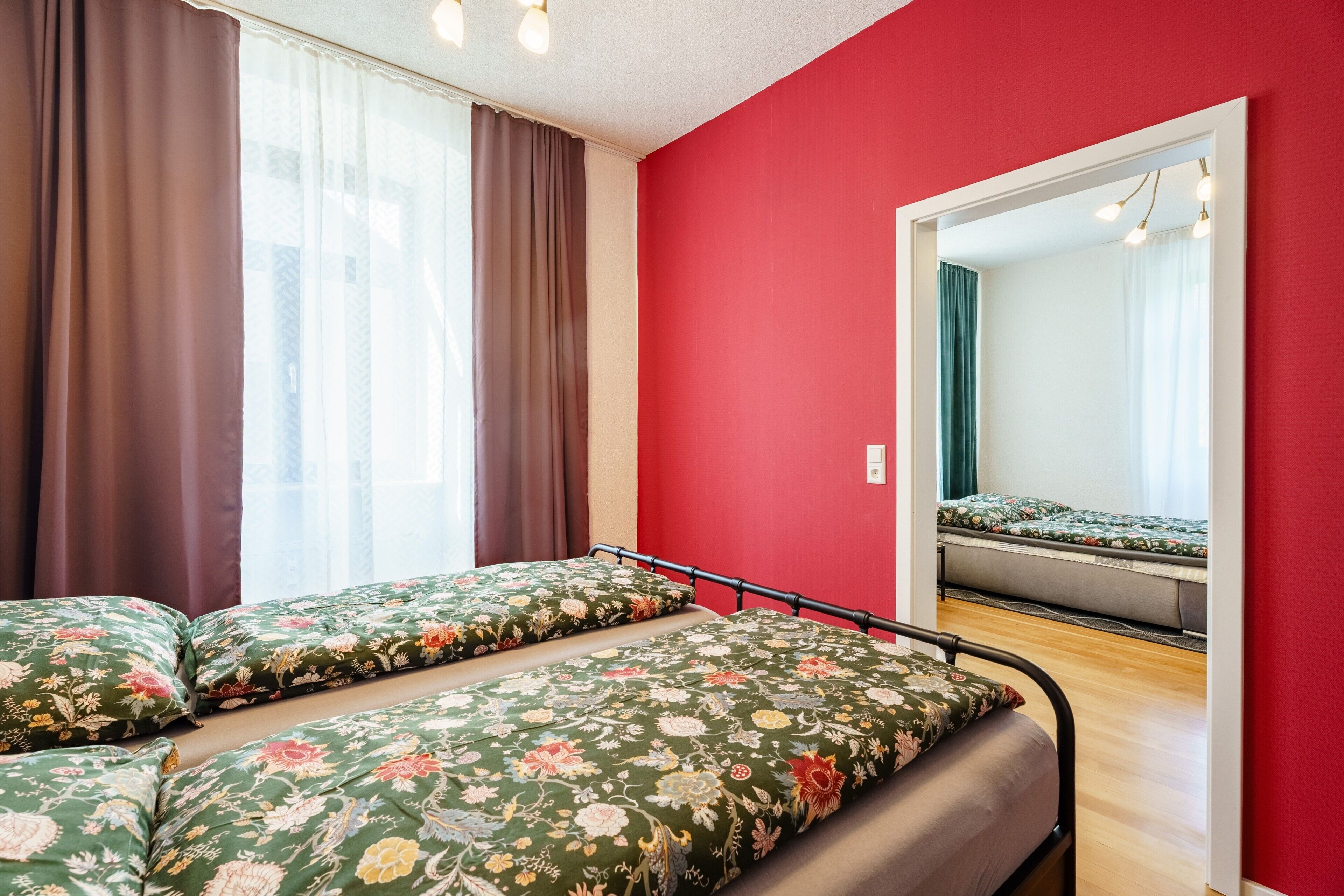 Une chambre avec deux lits, un mur rouge, un miroir et une fenêtre avec des rideaux.