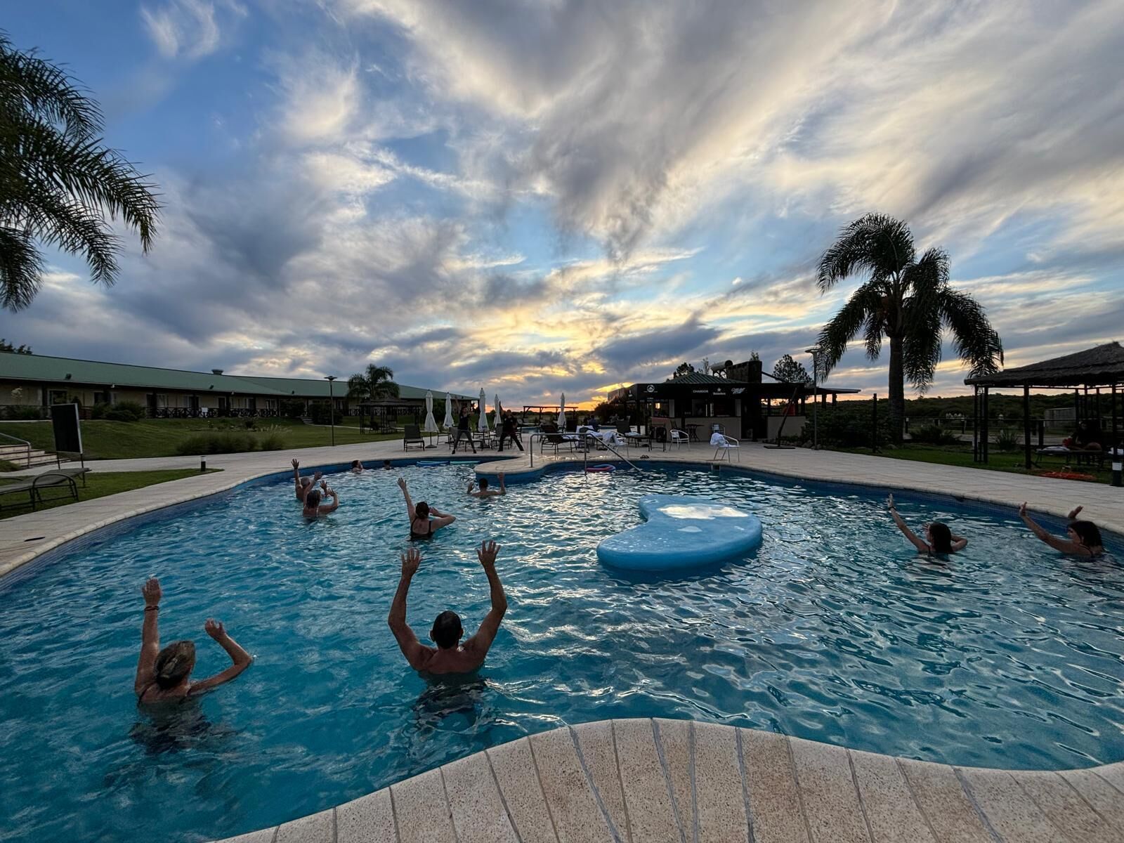 Une piscine où des personnes se baignent et profitent de l’eau, entourée de palmiers et d’un bâtiment doté d’un toit végétalisé.