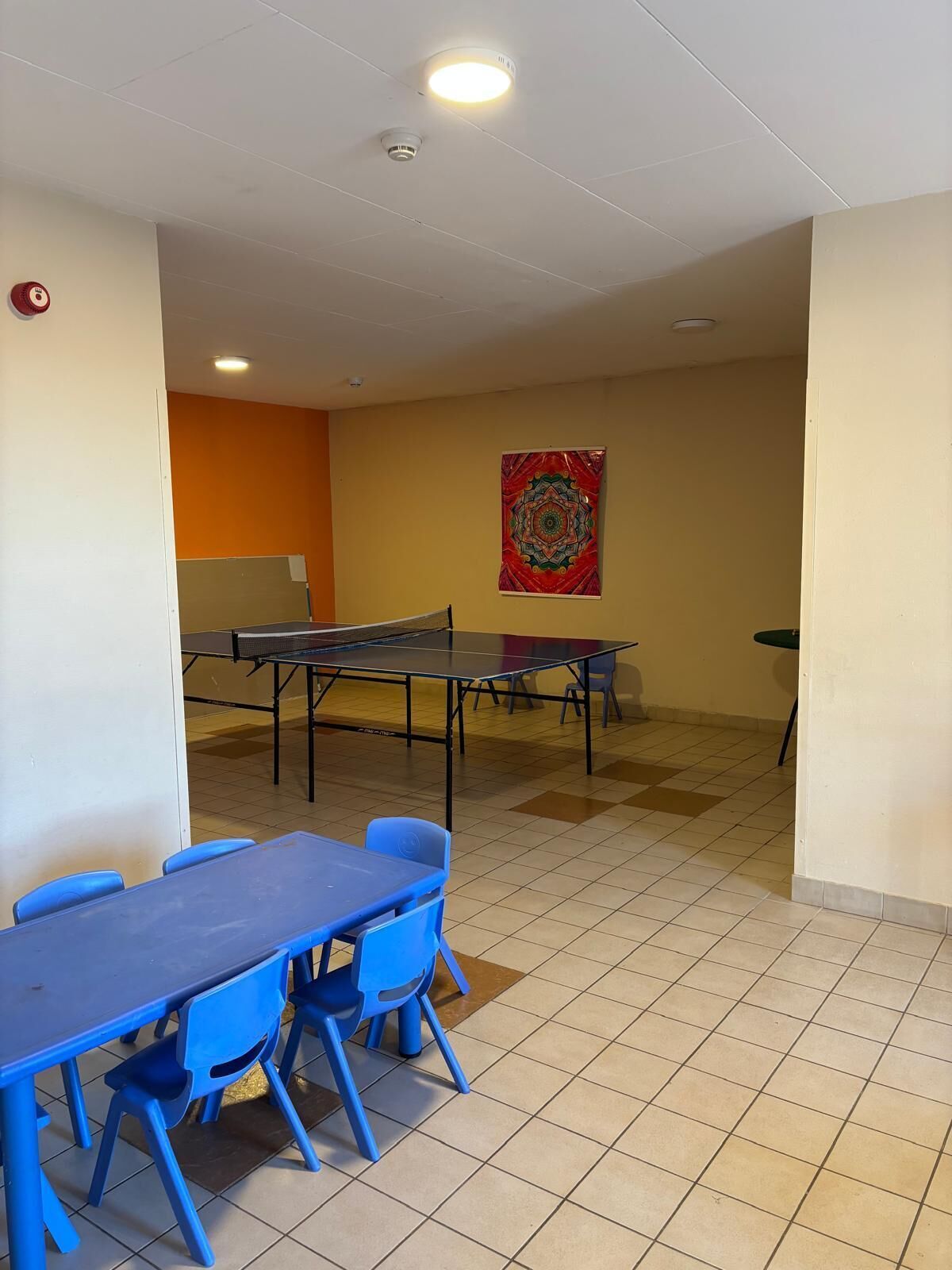 Una stanza con un tavolo da ping pong, sedie blu, una lavagna e un arazzo colorato appeso al muro.
