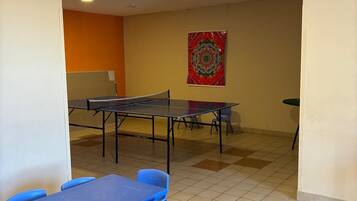 Una stanza con un tavolo da ping pong, sedie blu, una lavagna e un arazzo colorato appeso al muro.