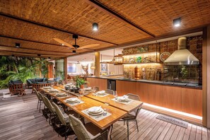 A spacious dining area with a long wooden table set for a meal, surrounded by wicker chairs. - Nana's House. Comfort and exclusivity in the best condominium in Praia do Forte. (MATA DO SÃO JOÃO)