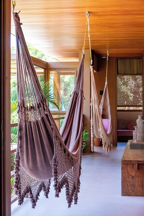 A room with two macramé hammocks, a wooden table, and a view of greenery outside. - Nana's House. Comfort and exclusivity in the best condominium in Praia do Forte. (MATA DO SÃO JOÃO)