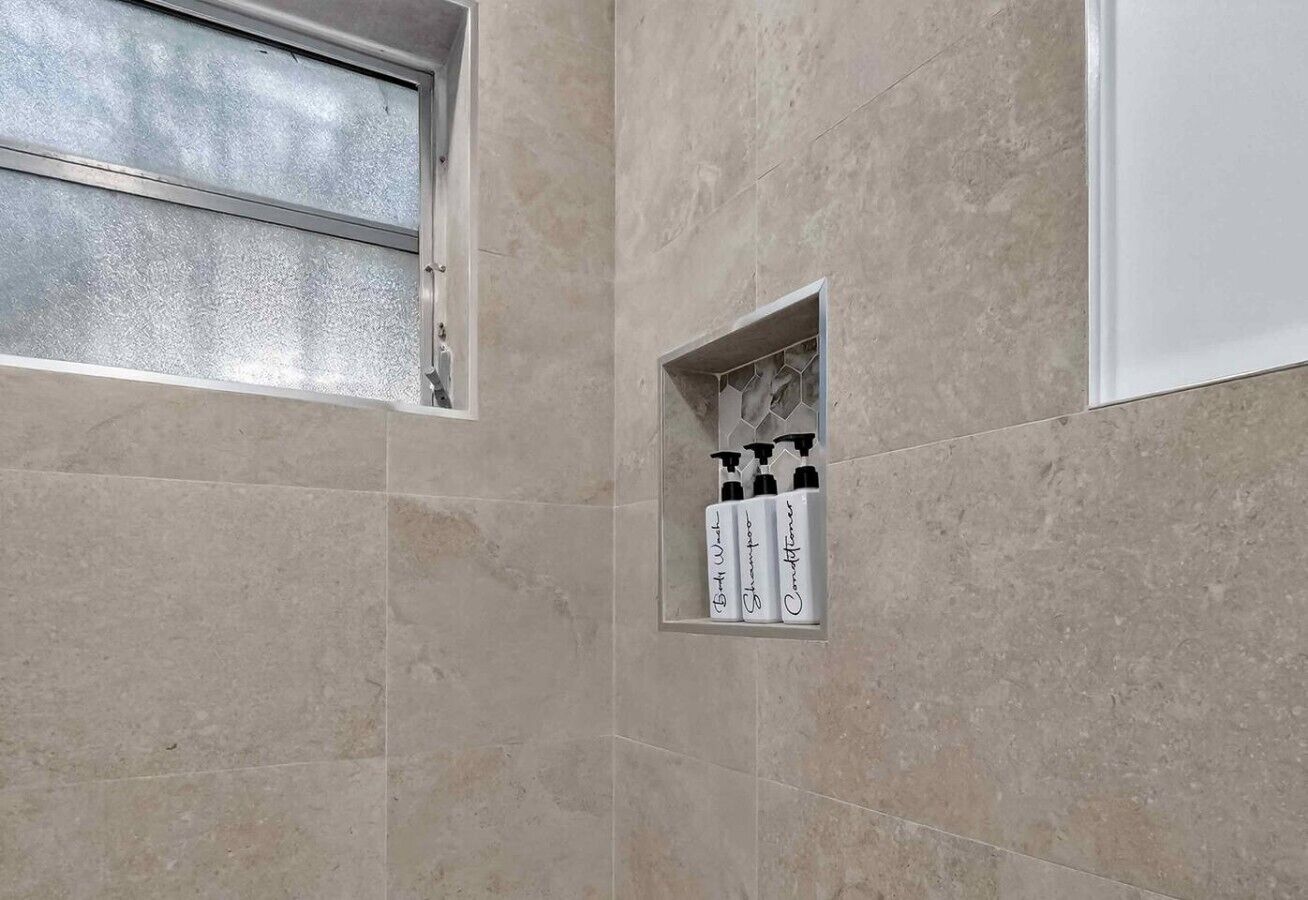 Um banheiro com azulejos bege, uma janela e um prateleira com artigos de higiene pessoal.