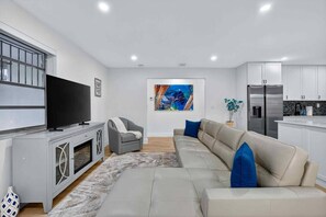 A modern living room with a sofa, a television, and a kitchen area with cabinets and a refrigerator. - Modern and Spacious 3BR - 2BA Miami - Free Parking (Miami)