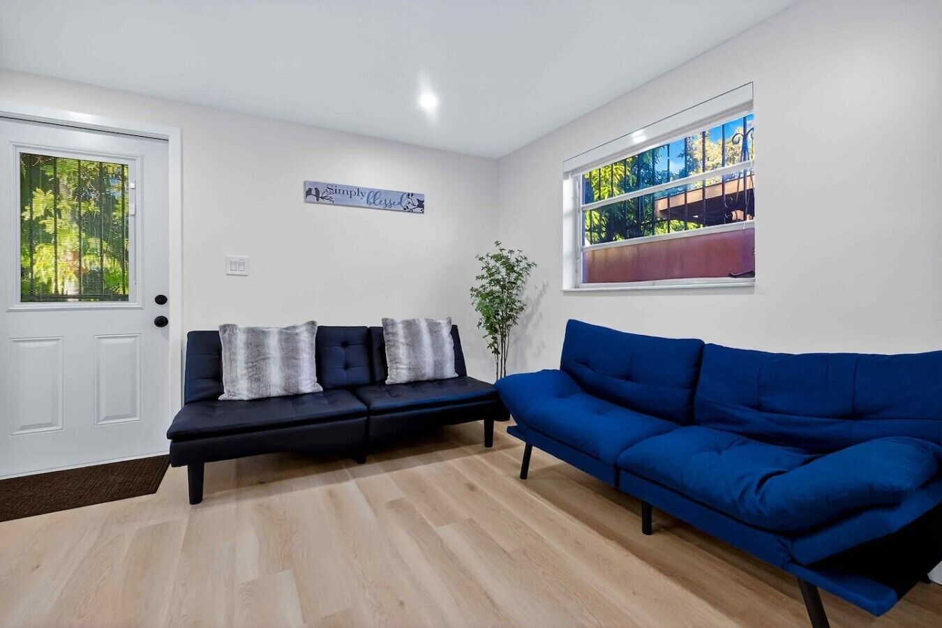 Een woonkamer met een donkerblauwe bank, een witte deur en een raam met uitzicht op bomen.