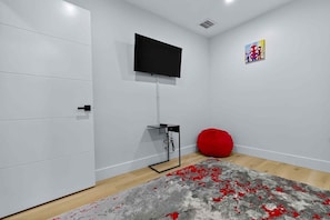 A modern room with a flat-screen TV mounted on the wall, a red bean bag chair, and a grey patterned rug on the floor. - Serenity Grove 3BR|2BA - Free Parking - Tropical (Miami)