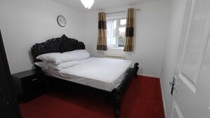 Une chambre à coucher avec un lit, une table de chevet, une horloge, une fenêtre avec des rideaux et un radiateur.