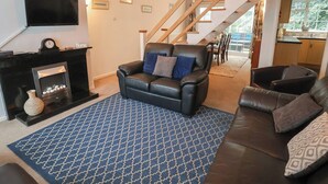 A living room with a fireplace, a sofa, a patterned rug, and a staircase leading to a kitchen. - Cromer Holiday Home (Cromer)