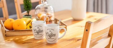Ein Holztisch mit zwei weißen Kaffeetassen, einem Glasbehälter, einer Topfpflanze und einer Schale Orangen.