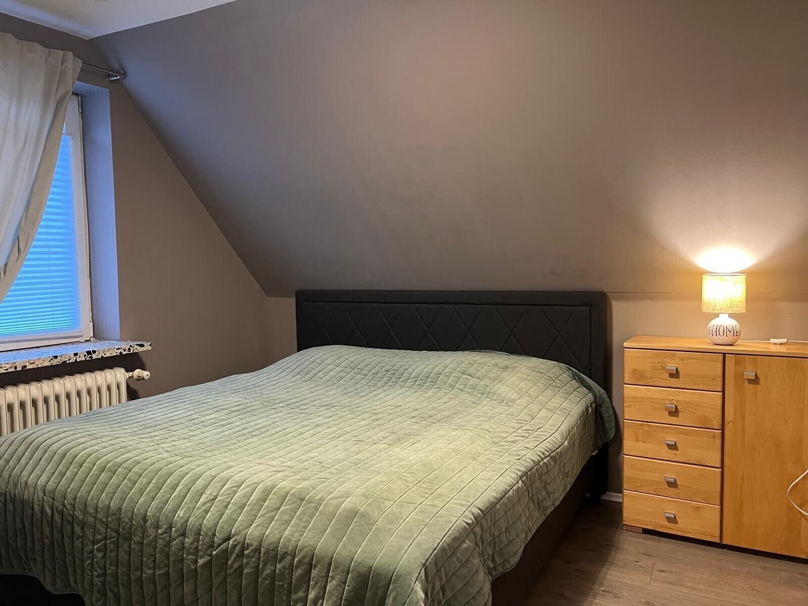 Et soveværelse med seng, et natbord med lampe, et vindue med persienner og en radiator.
