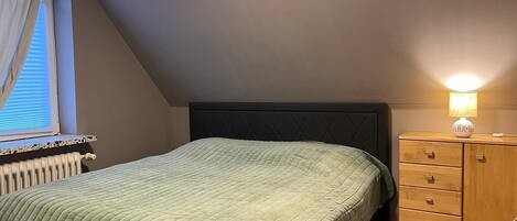 Et soveværelse med seng, et natbord med lampe, et vindue med persienner og en radiator.