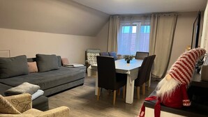 A modern living room with a sofa, a patterned armchair, a dining table with chairs, and a decorative gnome. - Apartment "Wohnung mit Fernblick" with Private Garden and Wi-Fi (Jübek)