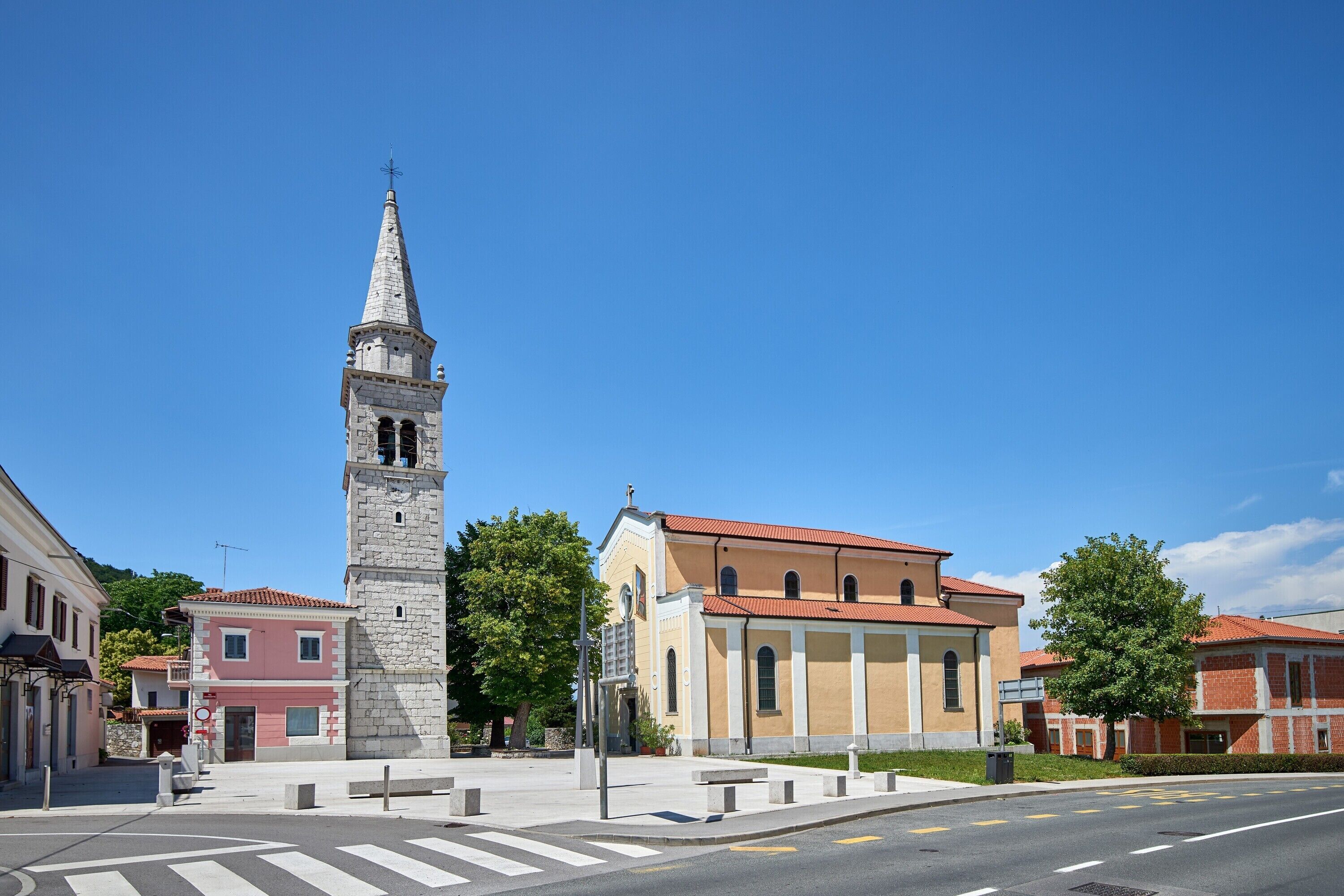 Een historisch kerkgebouw met een hoge klokkentoren, een gebouw met rode dakpannen en een zebrapad op een zonnig stadsplein.