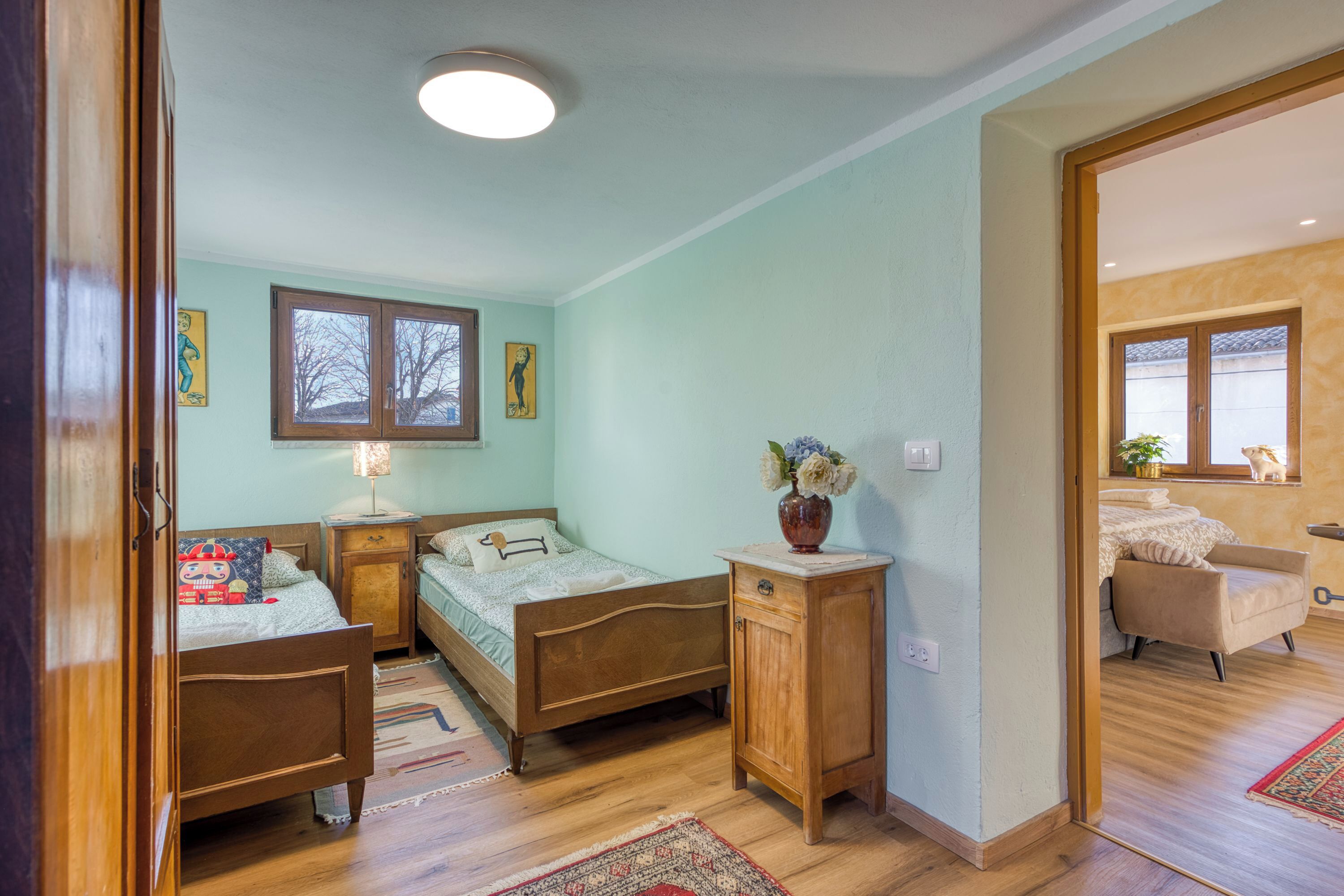 Une chambre à coucher avec deux lits en bois, une table de chevet, une commode, une fenêtre et une porte donnant sur une autre pièce.