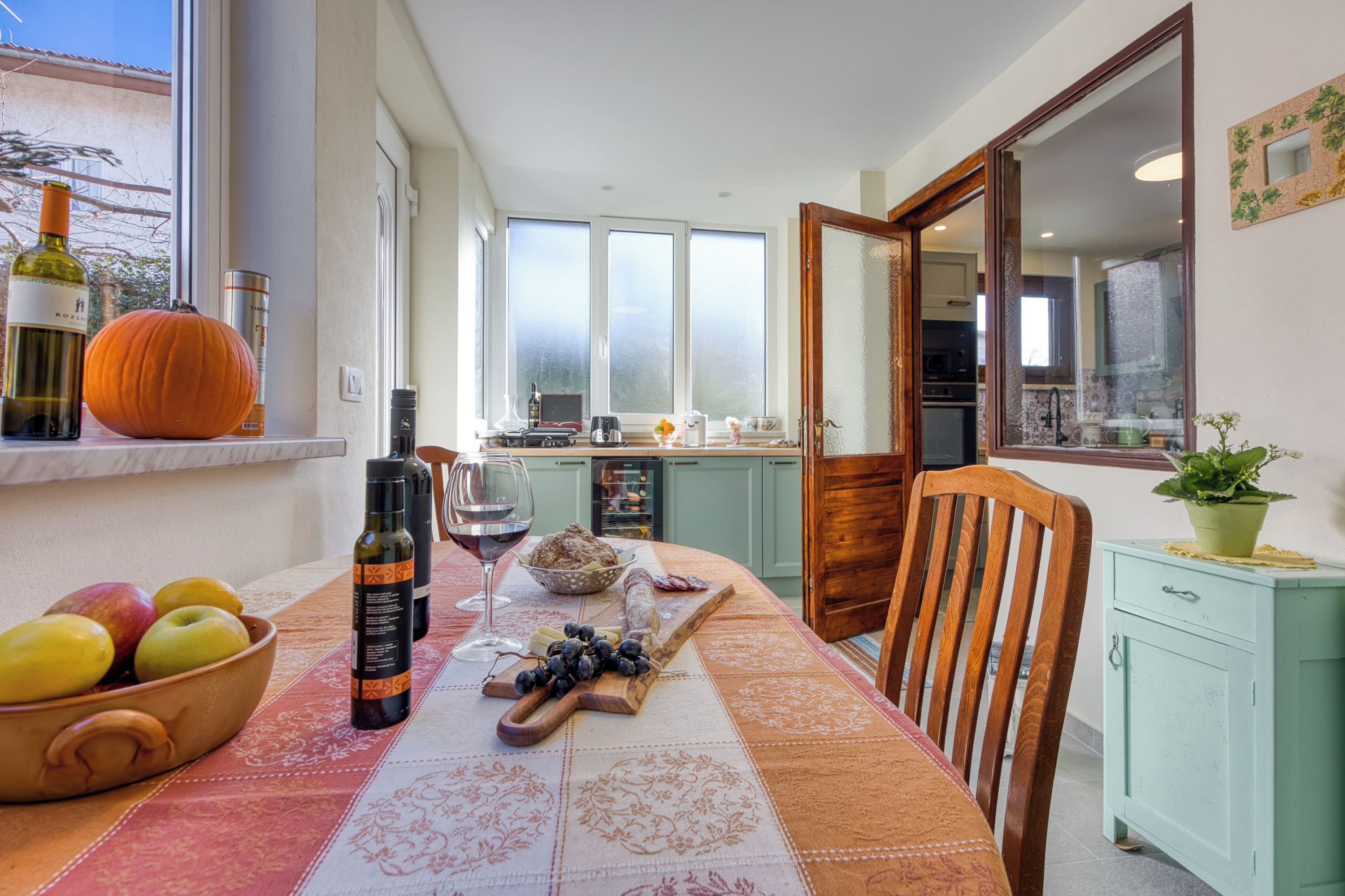 Un coin repas avec une table dressée pour deux personnes, agrémenté d’un saladier de fruits, d’une bouteille de vin et d’une planche à découper en bois sur laquelle sont disposés des raisins.