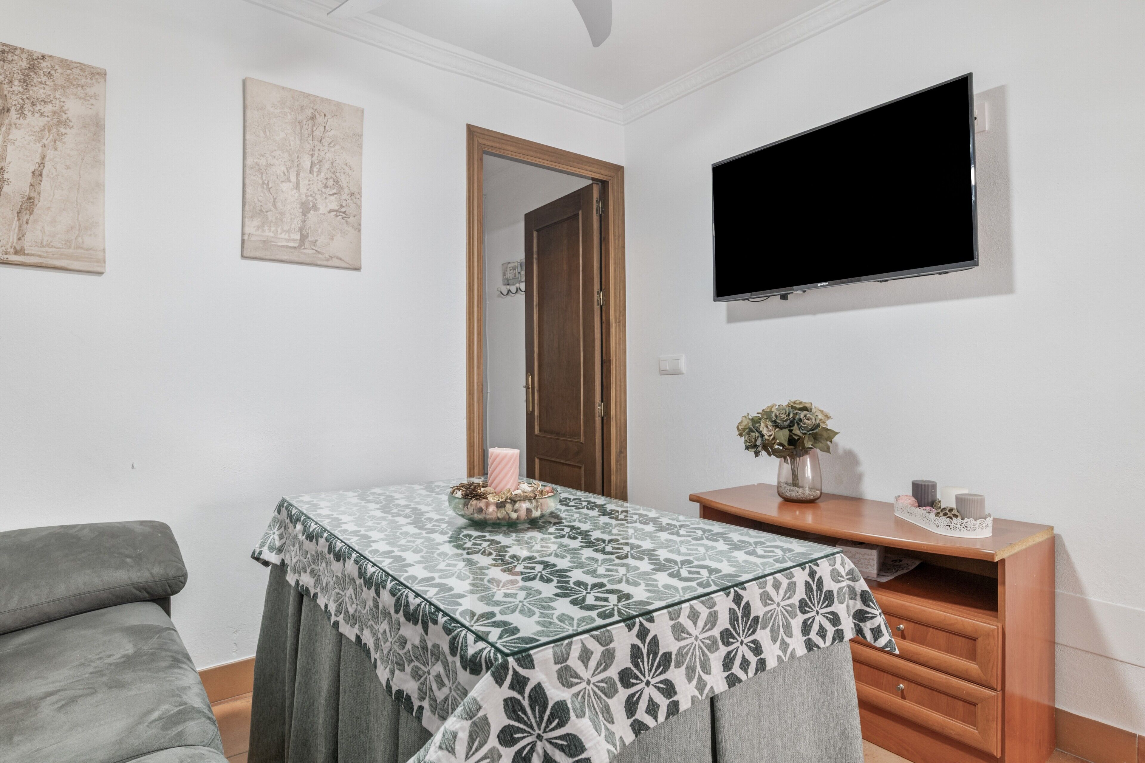 Une pièce avec un téléviseur fixé au mur, une table recouverte d’une nappe à motifs, un canapé et un meuble en bois agrémenté d’objets décoratifs.