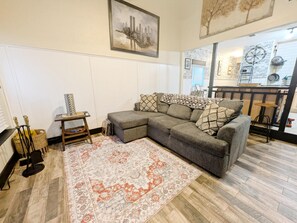 En stue med en hjørnesofa, et mønstret tæppe, et lille bord med pynteting og et bybillede på væggen.