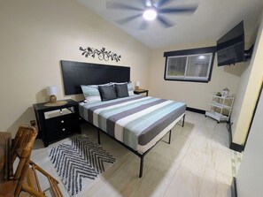 Een slaapkamer met een bed, nachtkastjes, een decoratief wandobject, een raam met jaloezieën, een plafondventilator en een witte schapunit.