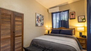 Un dormitorio con una cama, dos mesitas de noche, un ventanal con cortinas, un equipo de aire acondicionado y una puerta con manija.
