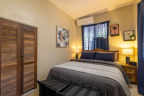 A bedroom with a bed, two bedside tables, a window with curtains, an air conditioning unit, and a door with a handle. - Coqui Haven Hideaway Rainforest View (luquillo)
