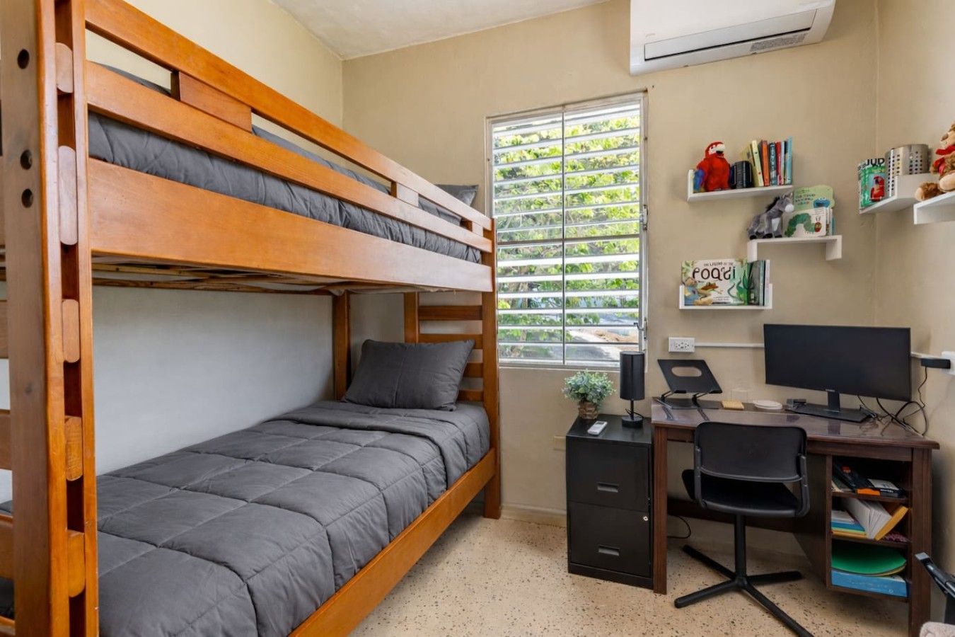 Una cama individual sobre otra con escritorio y silla, una ventana con persianas, y estantes con libros y objetos decorativos.
