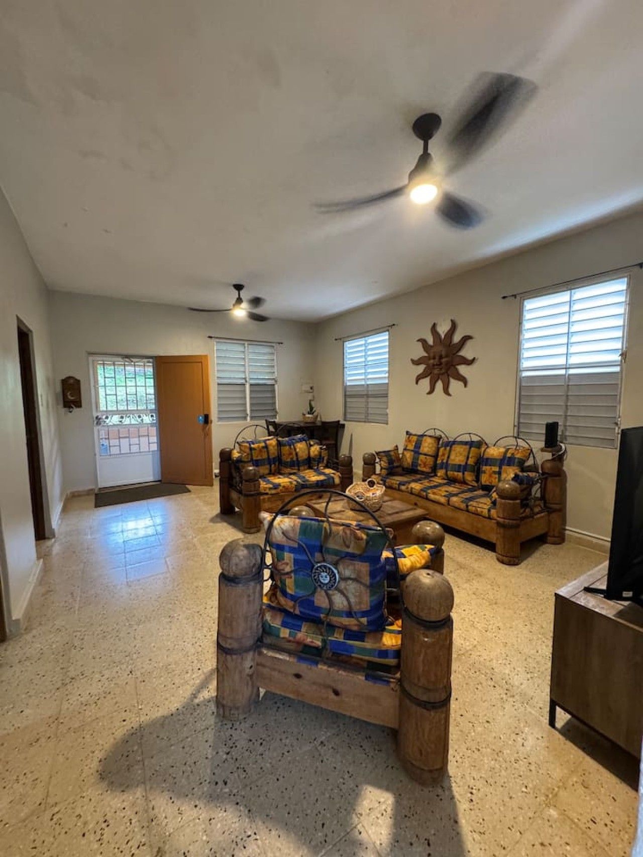 A spacious living room with a high ceiling, two ceiling fans, a large window with blinds, a wooden coffee table, and a television.