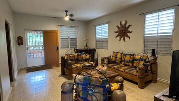A spacious living room with a high ceiling, two ceiling fans, a large window with blinds, a wooden coffee table, and a television.