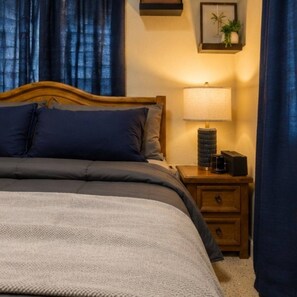 A neatly made bed with a grey and blue duvet, a bedside lamp, and a small wooden nightstand with a plant and a speaker. - Private 7-Acre El Yunque Retreat | Generator (luquillo)